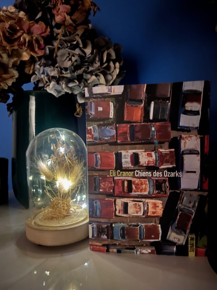 Photo du livre « Chiens des Ozarks » d’Eli Cranor posé près d’une lampe sous verre éclairée, devant un vase avec des fleurs, sur un fond bleu.